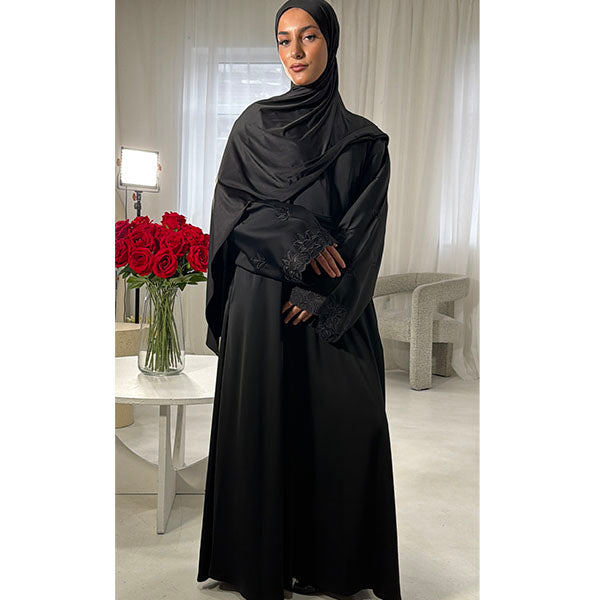 A woman wearing a long black abaya with matching black hijab stands indoors in a bright room with white curtains, a small table with red roses, and a light-colored chair in the background.