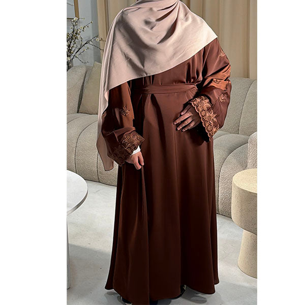 A woman wearing a long brown abaya with lace-trimmed sleeves and a matching headscarf, standing in a modern living room setting.