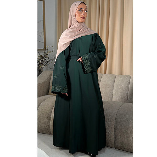 A woman wearing a dark green abaya with floral lace cuffs and a light beige hijab, standing in front of a beige sofa and curtains.