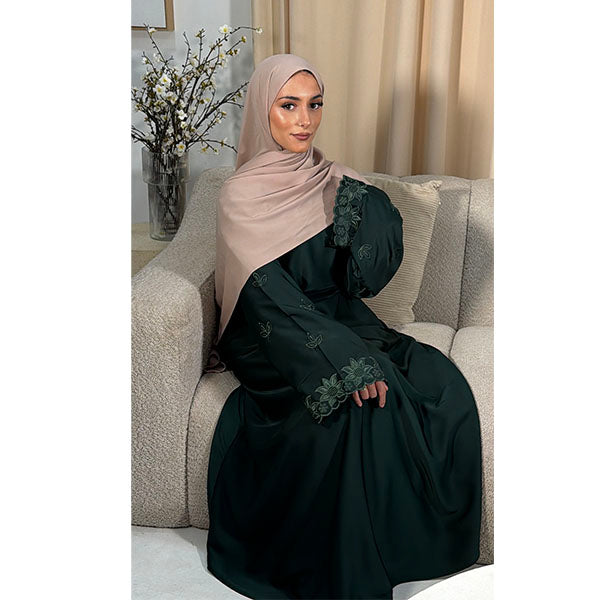 A woman wearing a dark green abaya with floral lace trim and a beige hijab, sitting on a textured beige sofa in a living room setting.